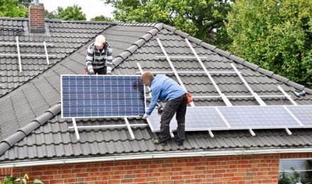 Dwóch mężczyzn na dachu domu jednorodzinnego, montujących panele fotowoltaiczne, zapewniające opłacalny prąd.