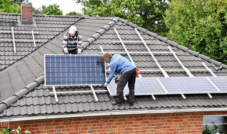 Dwóch mężczyzn na dachu domu jednorodzinnego, montujących panele fotowoltaiczne, zapewniające opłacalny prąd.
