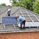 Dwóch mężczyzn na dachu domu jednorodzinnego, montujących panele fotowoltaiczne, zapewniające opłacalny prąd.